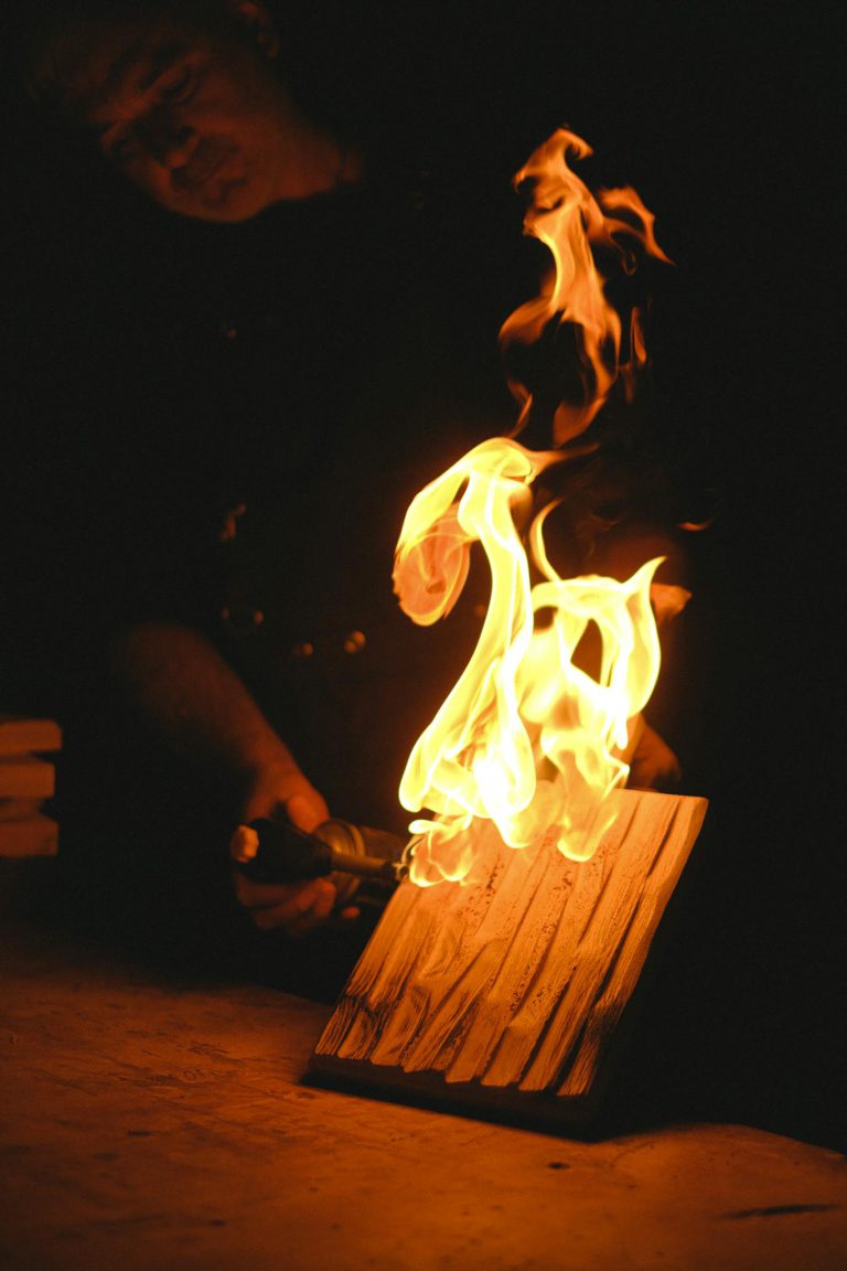 Anonymous male master with gas burner burning wooden plank while standing at table in dark carpentry with flame during work