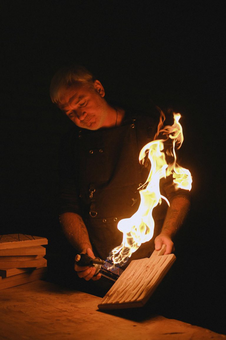 Concentrated male burning wooden detail with gas burner while working in dark studio with bright flame and stack of wood